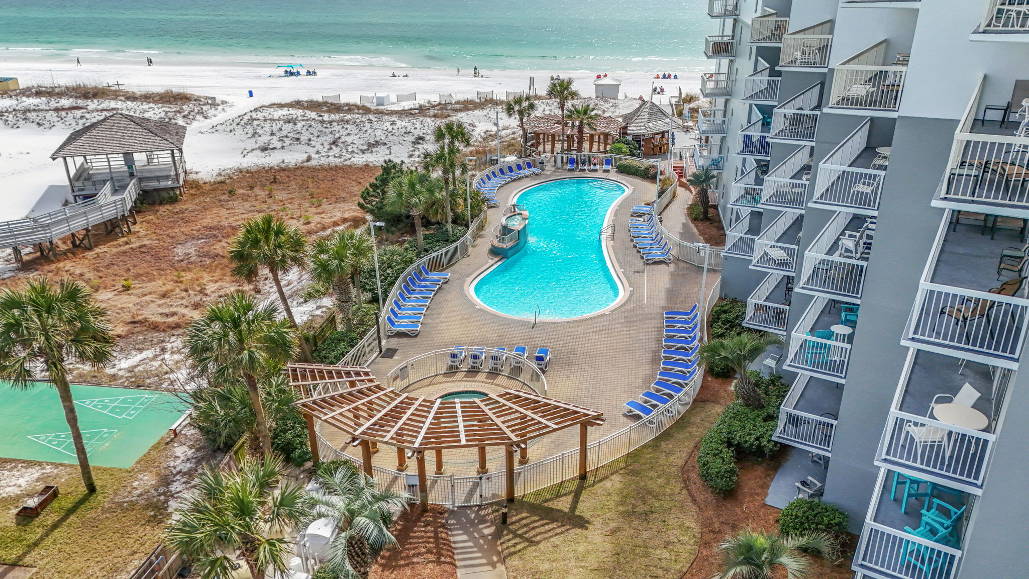 Aerial view of a beachfront hotel with a curvy outdoor pool, loungers, and wooden gazebo. The beach and ocean are visible in the background.