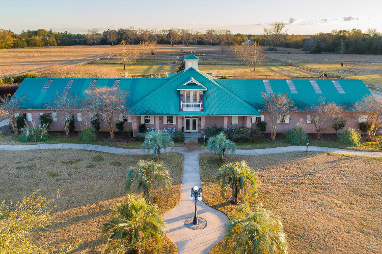 Aerial view of a large, symmetrical building with a green roof in a rural setting, surrounded by fields and trees. A pathway leads to the entrance, intersecting a landscaped area with palm trees.