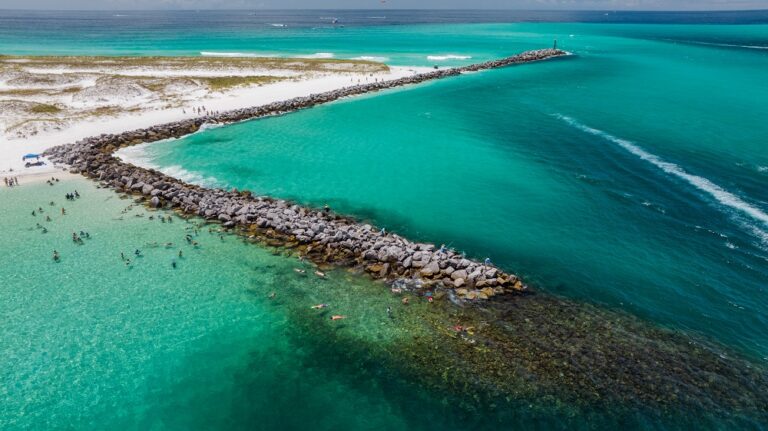 Aerial view of a rocky jetty extending into turquoise sea, with a sandy beach on the left and people swimming near the shore.