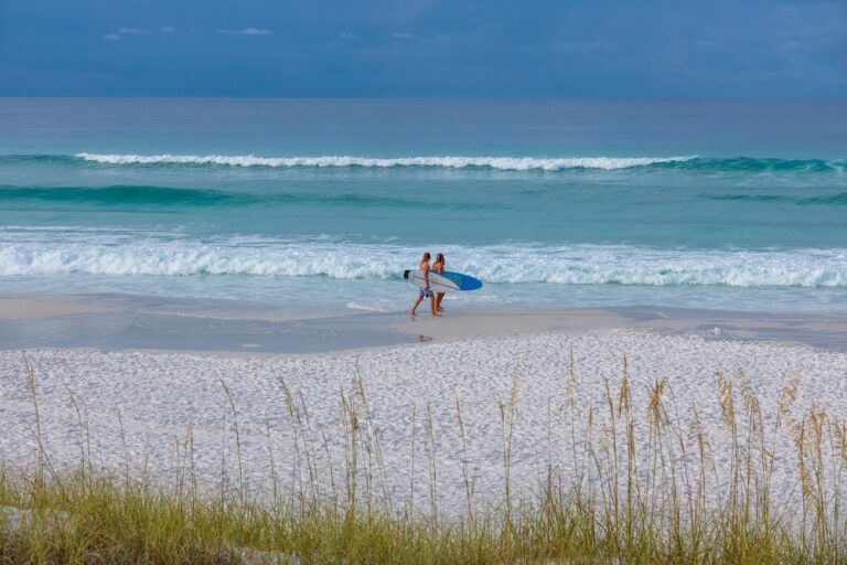Two people walk along a sandy beach carrying a surfboard, with waves rolling in under a cloudy blue sky.