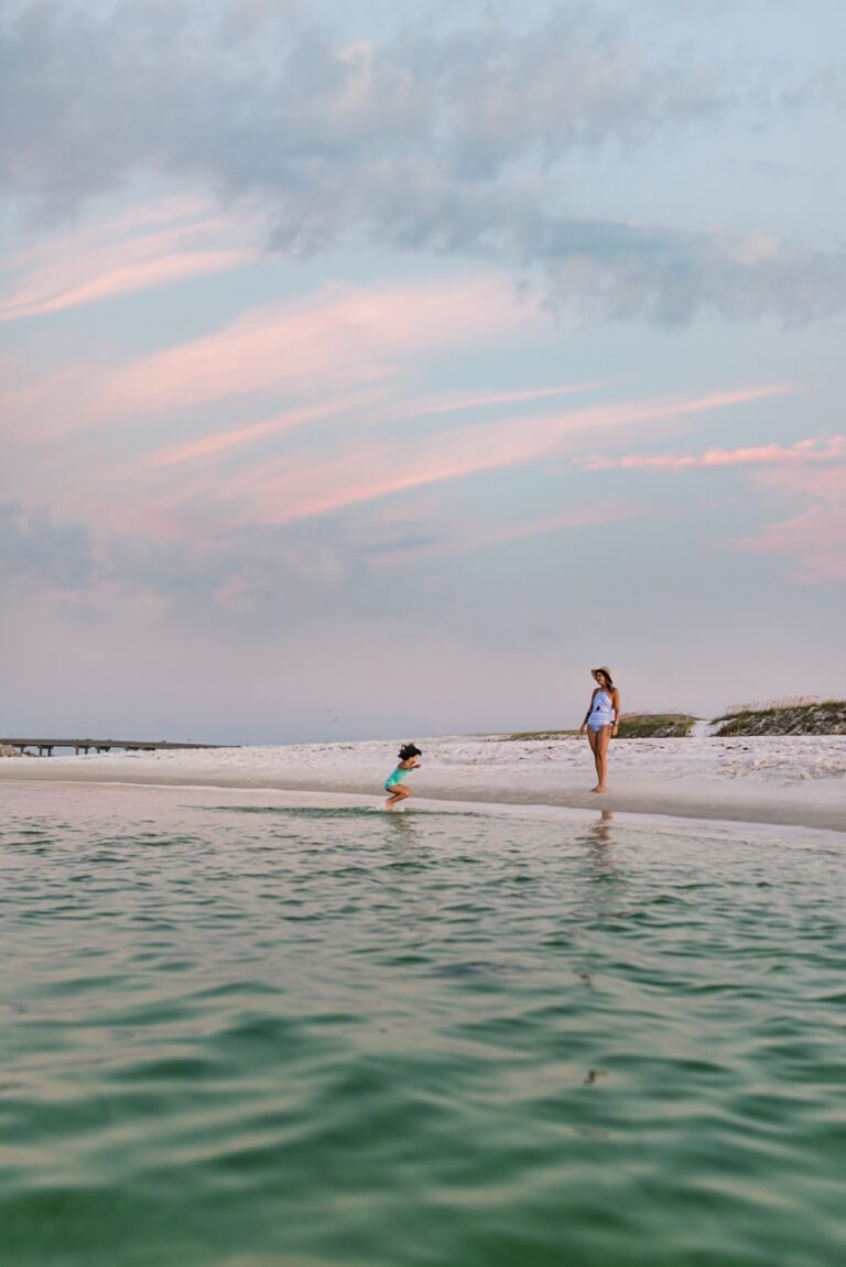 Two people stand near the shoreline of a beach under a sky with pink clouds. One is closer to the water, the other near the sand.