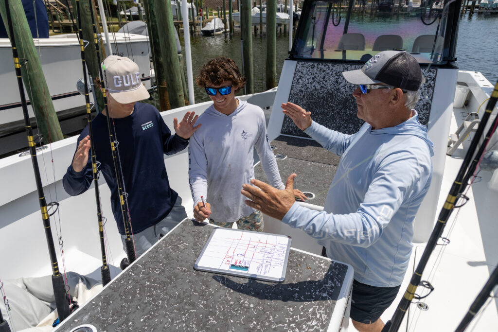 Three men on a fishing boat discuss a strategy using a whiteboard with a grid and notes; fishing rods are visible in the background.