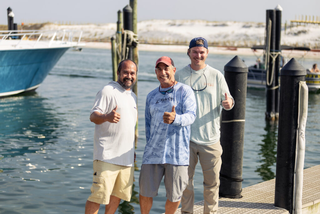 Three men stand on a dock by the water, smiling and giving thumbs up, with boats and sandy dunes visible in the background.