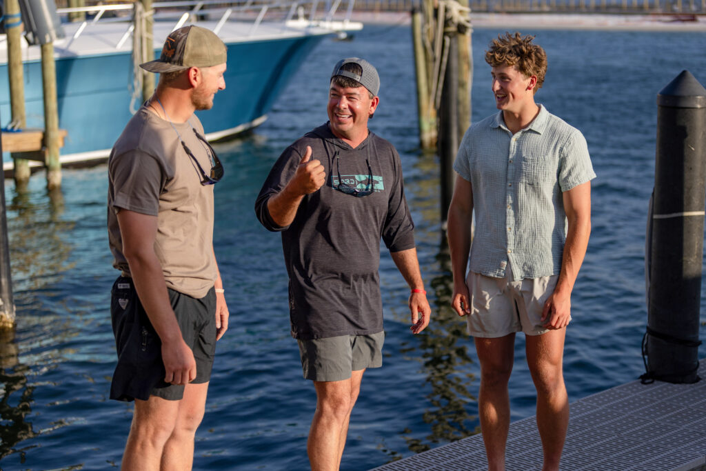 Three men stand and converse on a dock by the water, with a boat and wooden posts in the background on a sunny day.