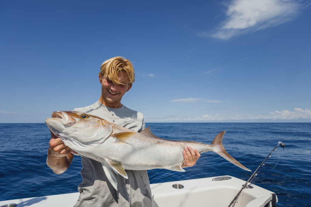 Person holding a large fish on a boat with a fishing rod in the background, under a clear blue sky.