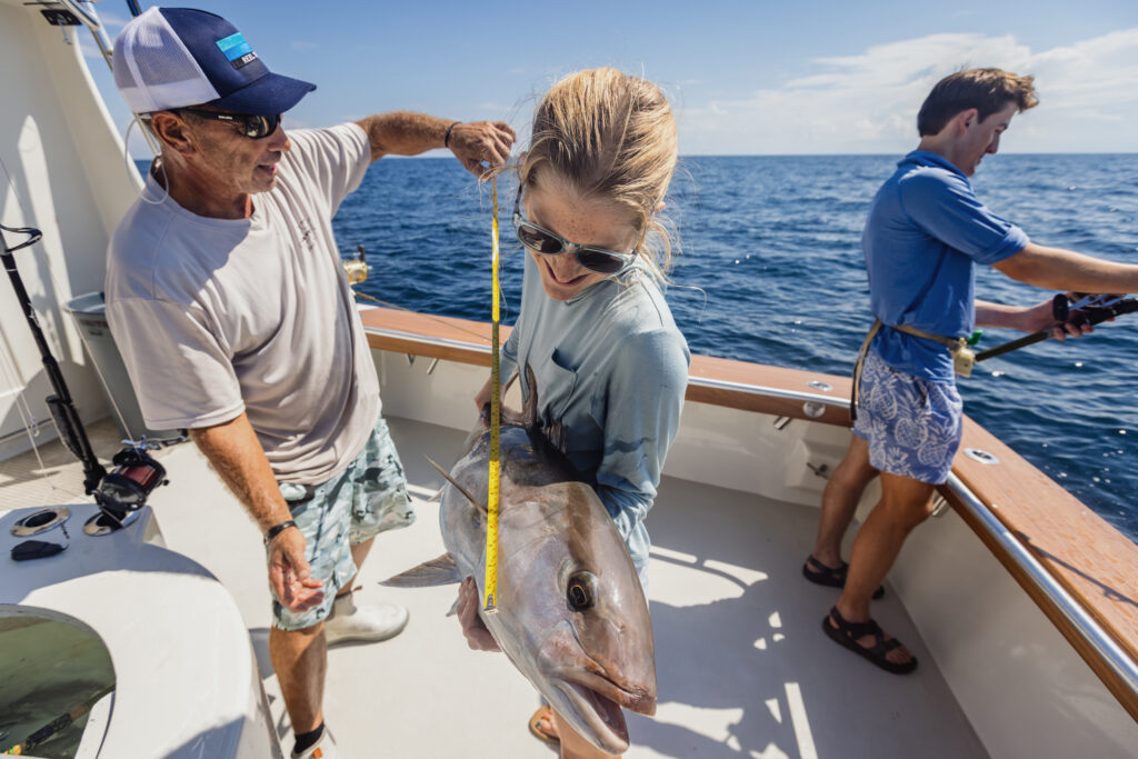 Two people measure a fish on a boat against an ocean backdrop. Another person is fishing in the background.