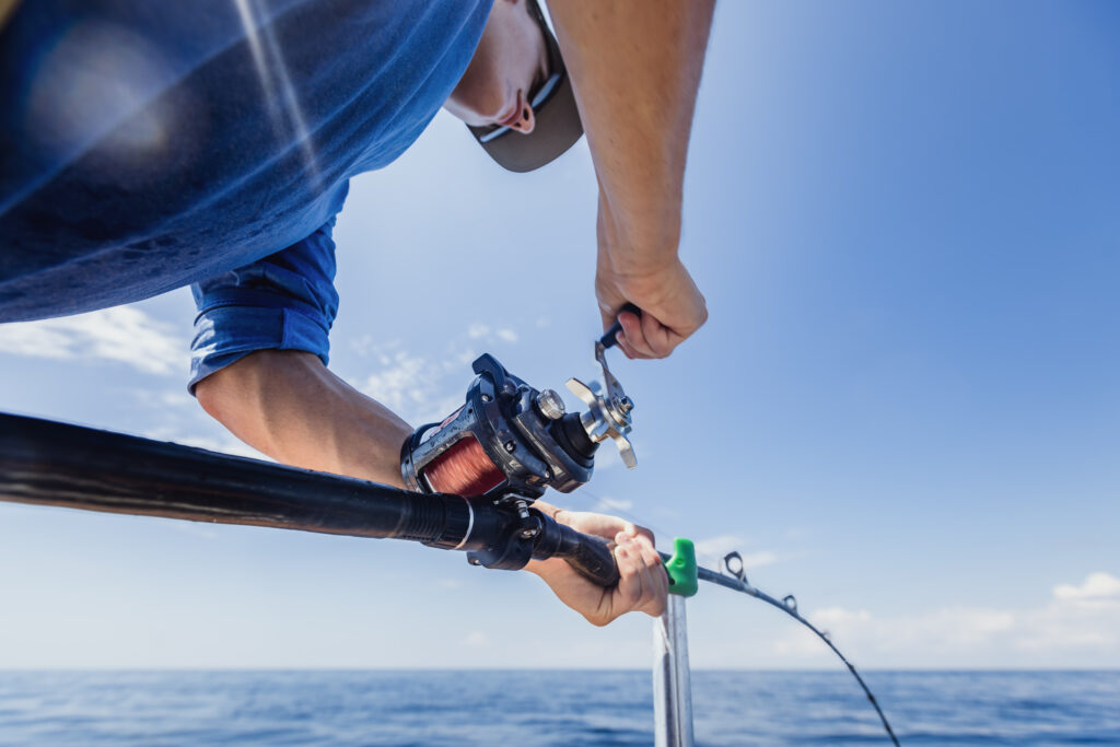 Person in a blue shirt reeling in a fish on a fishing rod over a calm ocean under a clear blue sky.