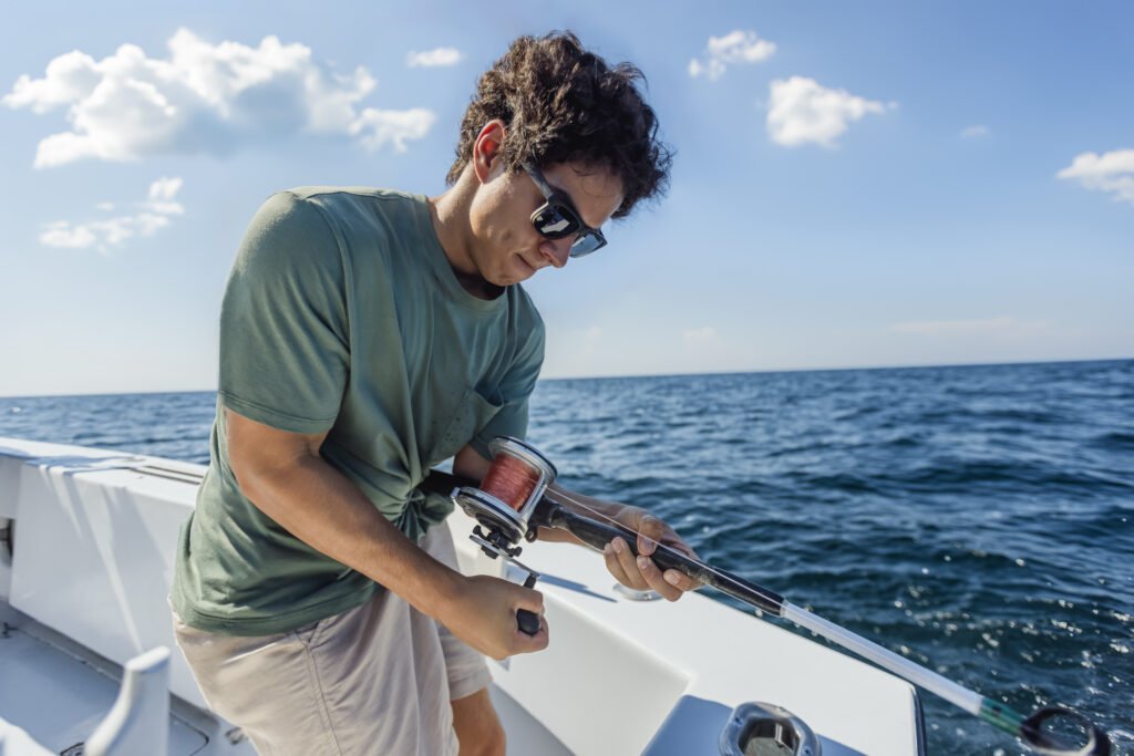 Person in a green shirt and sunglasses is operating a fishing rod on a boat under a clear blue sky.