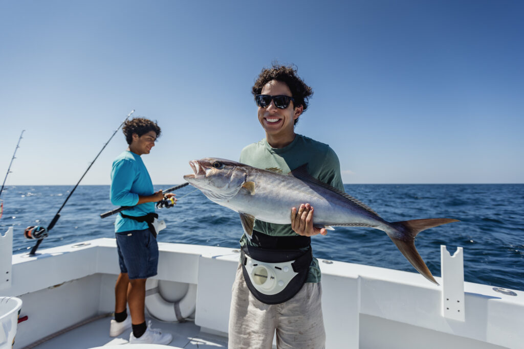 Person holding a large fish on a boat with another person in the background; both are smiling under a clear blue sky.