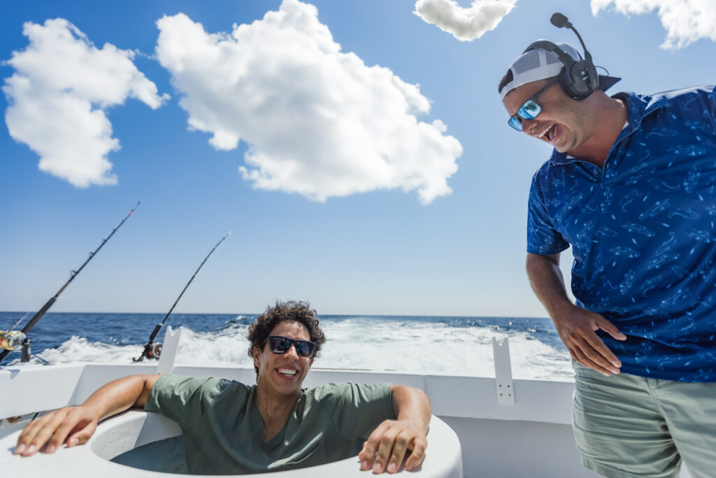 Two men on a boat under a cloudy sky, one sitting in a circular seat and smiling, the other standing with a headset and cap, both appear relaxed, with fishing rods in the background.