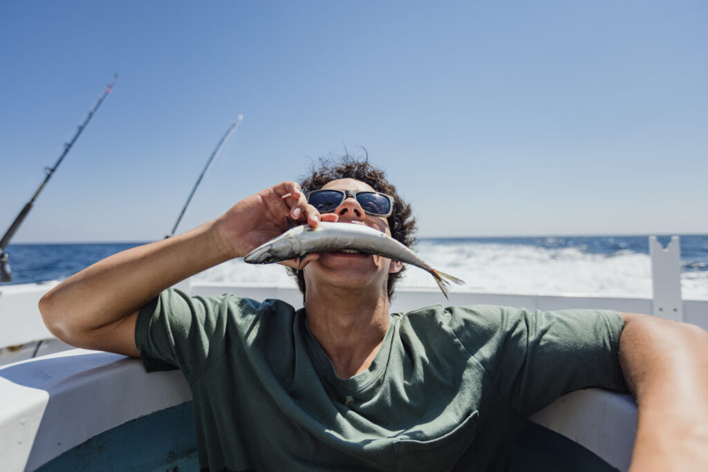 Person on a boat playfully pretending to eat a fish, with sunglasses and a green shirt. Ocean and fishing rods in the background.