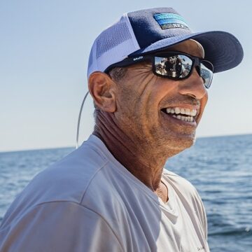 Man wearing a cap and sunglasses smiles outdoors, with the ocean in the background.