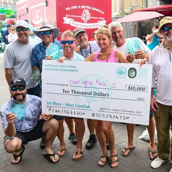 A group of eight people pose with a large novelty check for $10,000 at the Emerald Coast Open Lionfish Tournament. They are outdoors, in casual attire, standing in front of a red food truck.