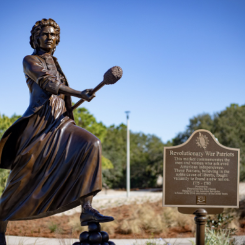 Bronze statue of a woman in historical attire holding a weapon, accompanied by a plaque honoring Revolutionary War patriots, set outdoors with trees and sky in the background.