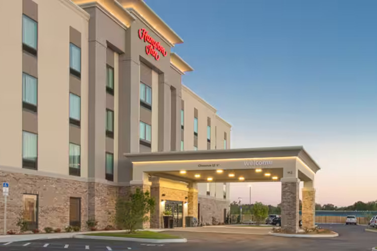 The image shows the exterior entrance of a modern Hampton Inn hotel at sunset with a covered driveway and visible signage.