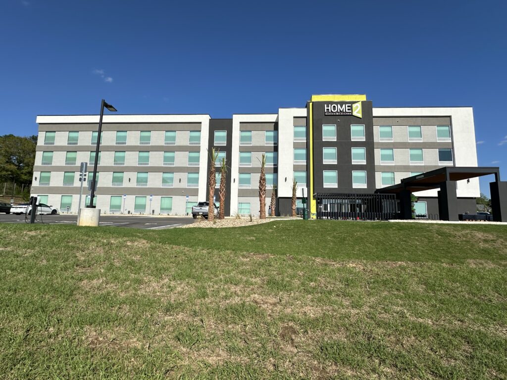 A four-story Home2 Suites by Hilton hotel with a flat roof, large windows, and a parking lot in front under a clear blue sky.