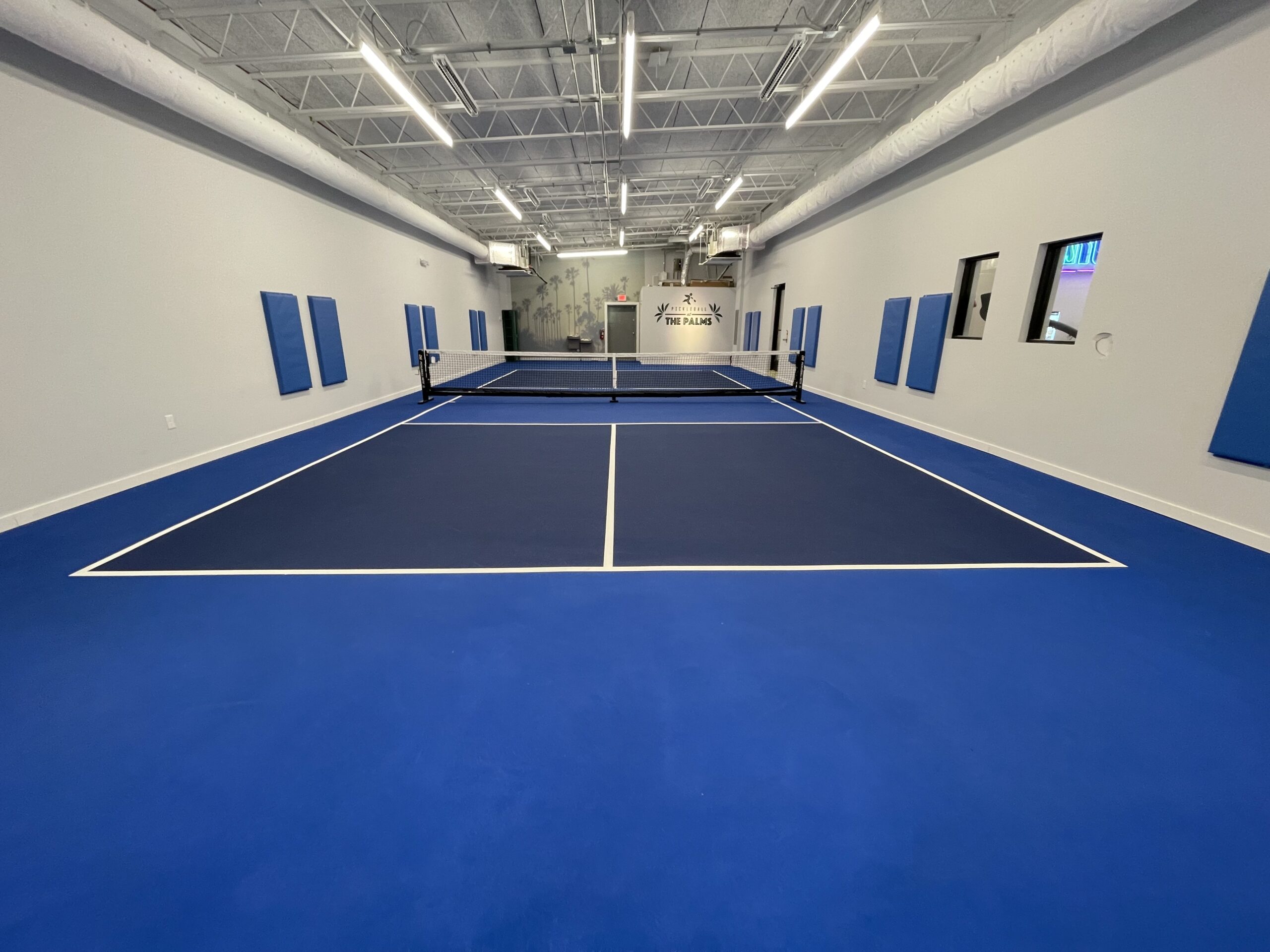 Indoor pickleball court with blue floor and walls, net in the center, and overhead lighting.