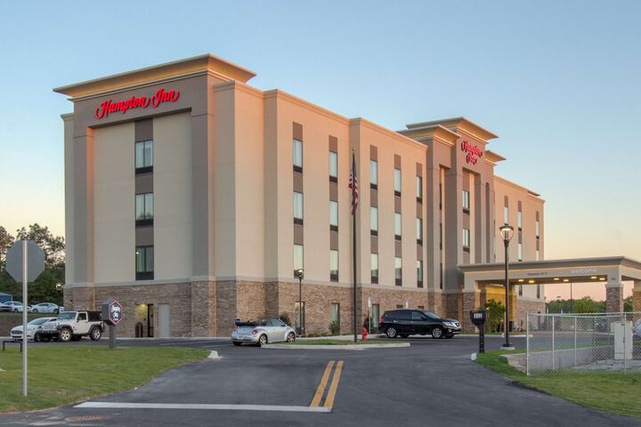 Exterior view of a four-story Hampton Inn hotel with cars parked in front and a driveway leading to the entrance.