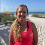 Woman with long blonde hair wearing a pink tank top stands on a sandy beach with the ocean and blue sky in the background.