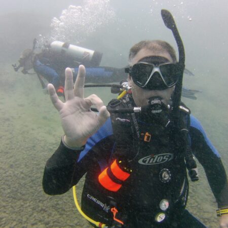 A scuba diver underwater gives an "OK" hand signal to the camera, with another diver visible in the background.