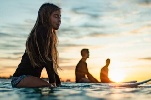 A young girl sits on a surfboard in the water at sunset, with two other people on surfboards in the background.