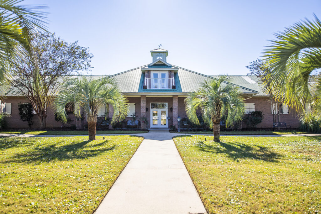 A single-story brick building with a green metal roof, central cupola, palm trees, and a concrete walkway leading to the entrance on a sunny day.