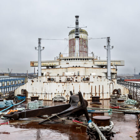 View of a large, rusted ship&rsquo;s bow with an anchor and ropes on deck, docked at an industrial port under a cloudy sky.
