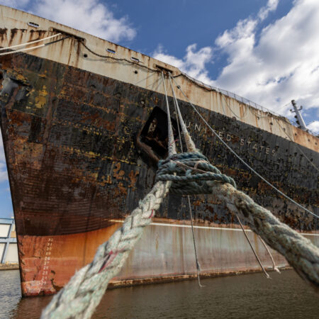 Close-up view of the bow of a large, rusted ship moored at a dock, with thick ropes securing it over the water.