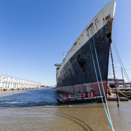 Large, rusted ship docked at an industrial port, with a small tugboat alongside and warehouses and power plant visible in the background under a clear blue sky.