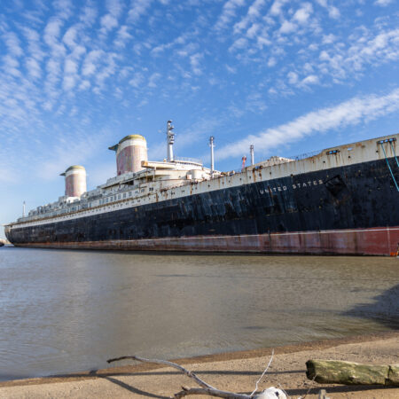 A large, rusting ship named &ldquo;United States&rdquo; is docked in a harbor under a blue sky with scattered clouds.