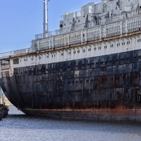 A large, rust-stained ship hull sits partially submerged in water at a dockyard, with industrial structures visible in the background.