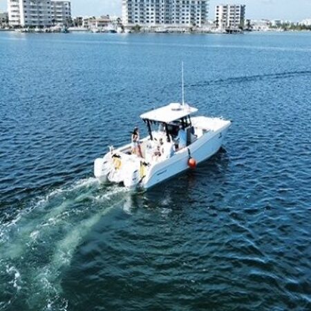 A white motorboat with several people on board moves through blue water near shoreline buildings under a clear sky.