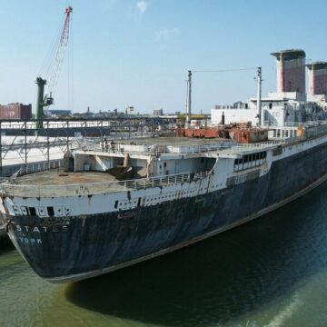A large, rusted ship named "United States" is docked in a harbor beside industrial buildings and cranes under a clear sky.