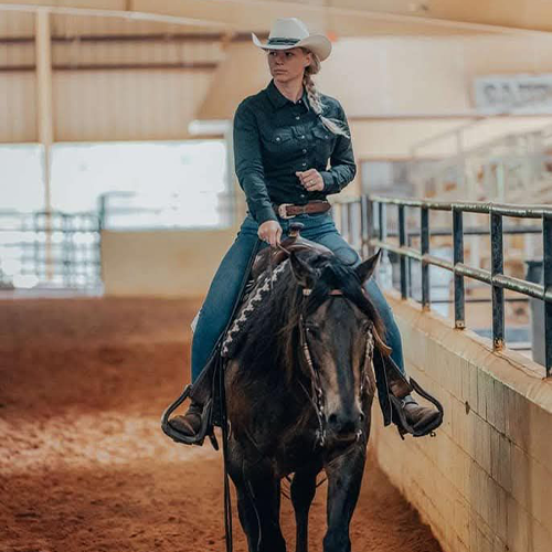 Person wearing a hat and denim attire riding a dark horse inside an indoor riding arena with sandy ground.