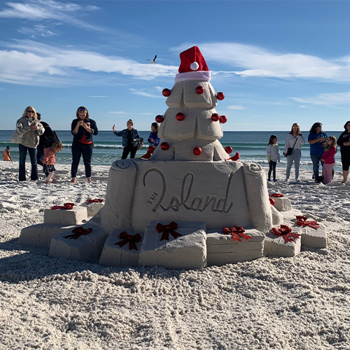 A sand sculpture decorated like a Christmas tree with red ornaments and a Santa hat, on a beach with people standing in the background.