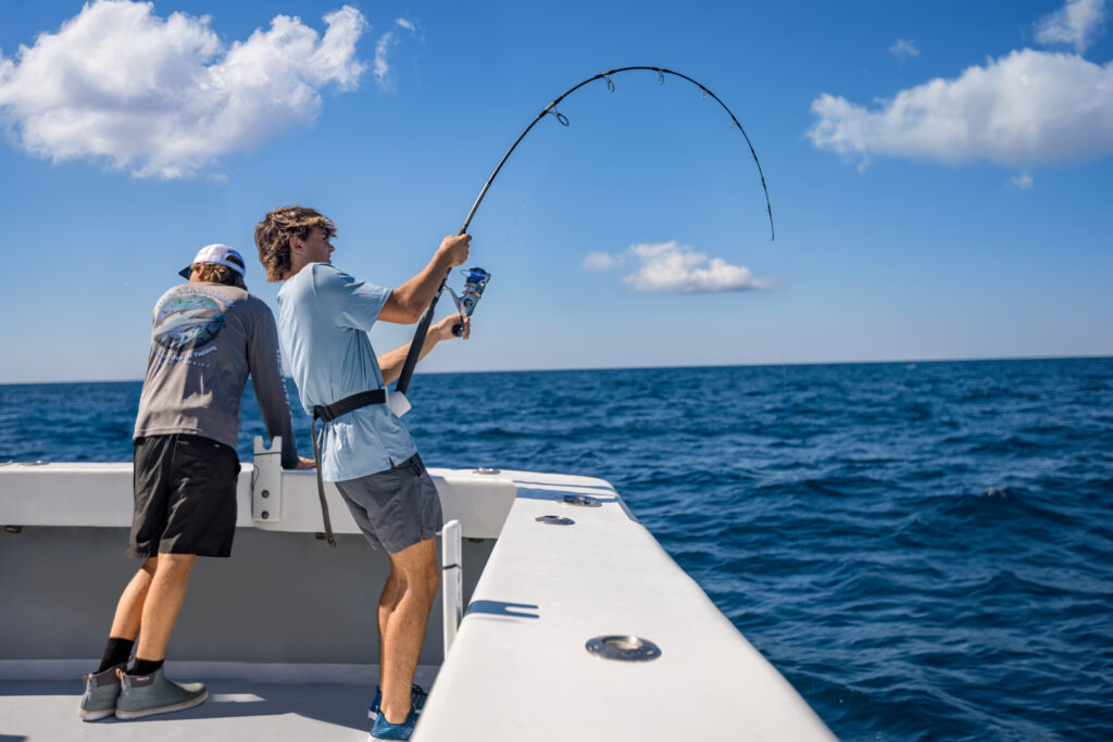 Two men stand on a boat in the ocean; one is reeling in a fishing rod, while the other watches. It is sunny with a few clouds in the sky.