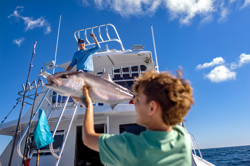 A boy holds up a large fish on a boat while a man stands on the upper deck, giving a thumbs up under a clear blue sky.