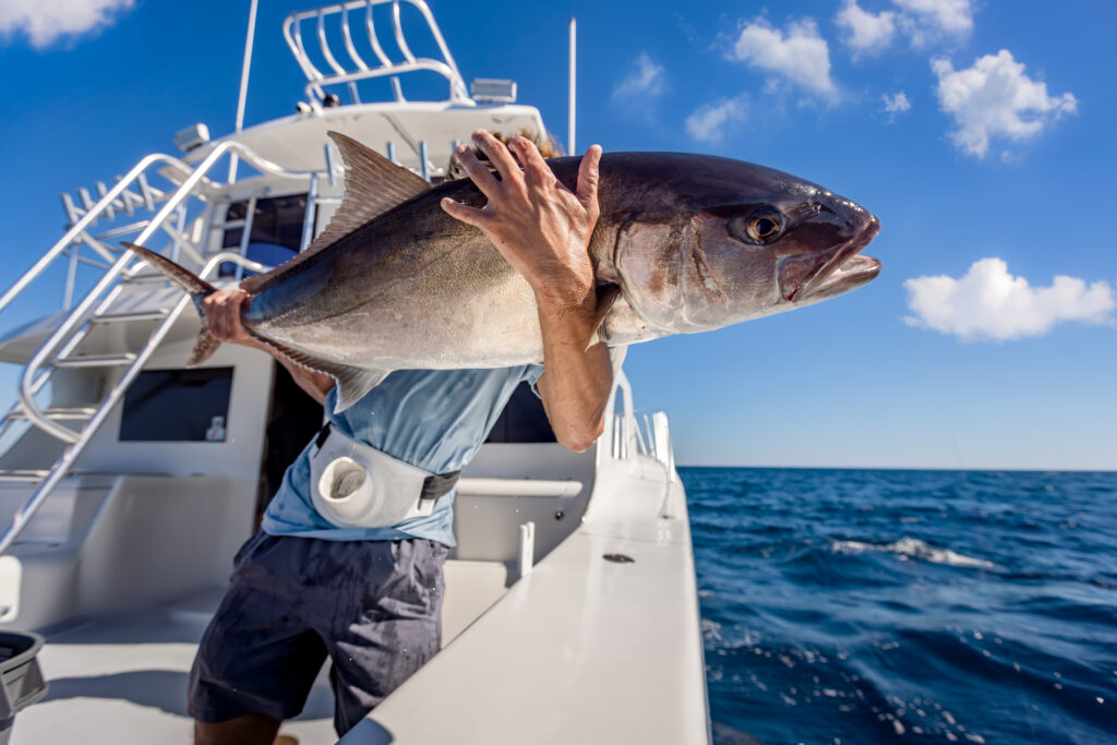 Person holding a large fish on a boat at sea under a bright blue sky with a few clouds.