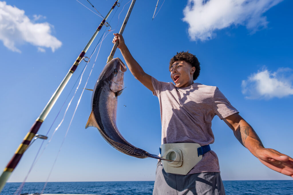 A person on a boat holds up a large fish caught on a fishing rod, wearing a fishing belt, with the ocean and blue sky in the background.