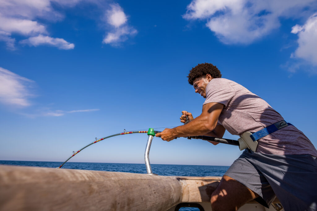 A person stands on a boat holding a bent fishing rod, appearing to reel in a catch under a bright blue sky.
