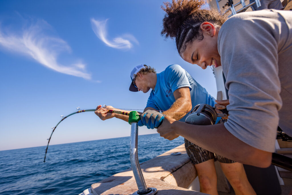 Two people on a boat work together to reel in a fish, one holding the fishing rod and the other assisting, with blue sky and ocean in the background.