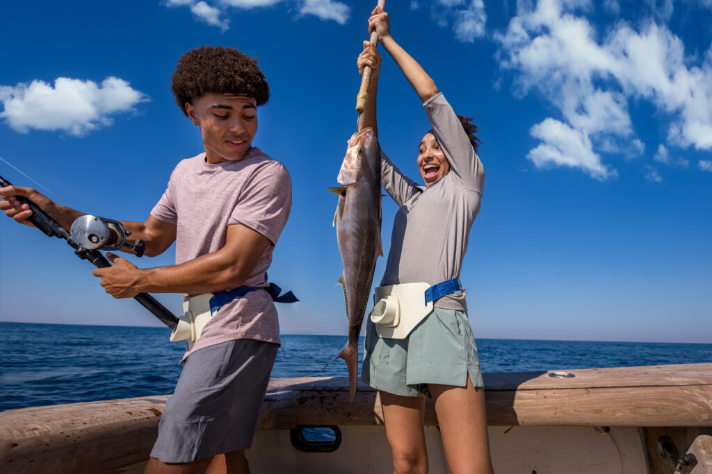 Two people on a boat, one holding a fishing rod and the other raising a large fish, with the ocean and blue sky in the background.