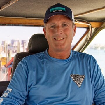 A man in a blue fishing shirt and cap sits inside a boat, smiling at the camera with a marina visible in the background.
