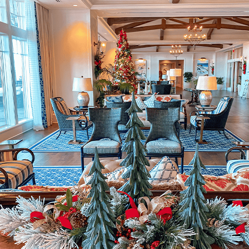 A decorated hotel lobby features a Christmas tree, festive wreaths, and small artificial pine trees on a table in the foreground. Blue rugs and seating areas fill the space.