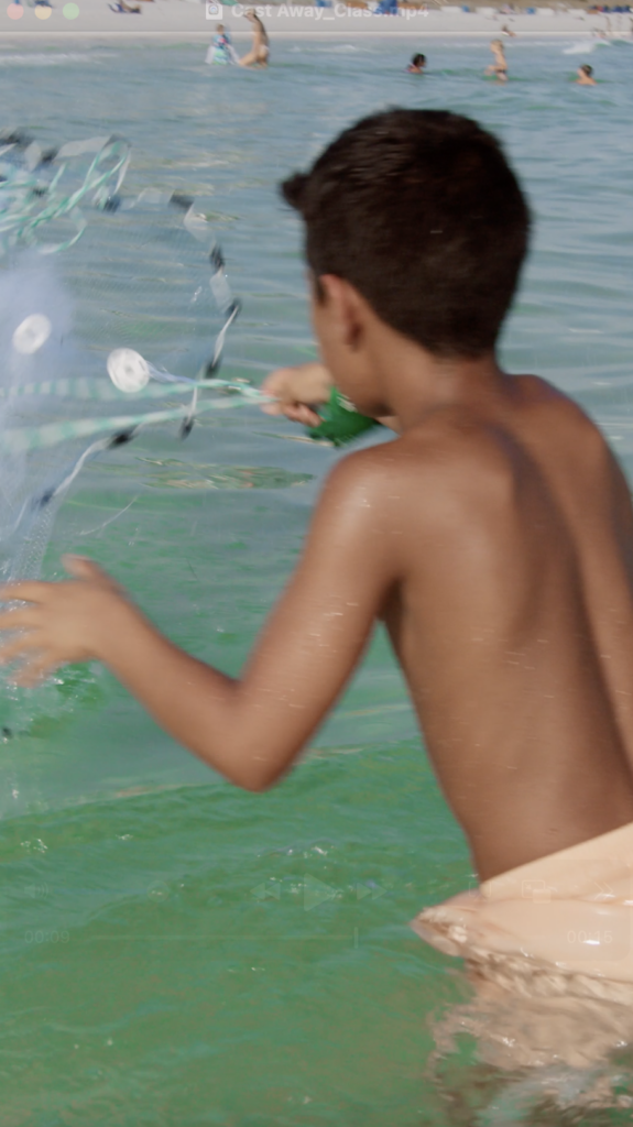 a young boy stands waist deep in the ocean and throws a fishing net