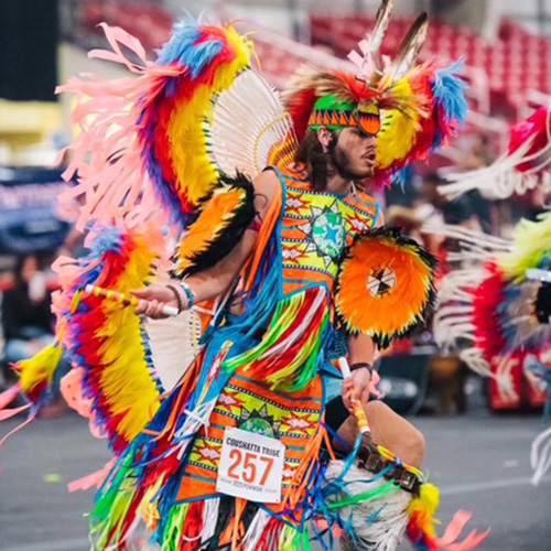 A person in a colorful, feathered Native American dance regalia performs at an indoor event, wearing the number 257.