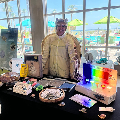 A person in a sea turtle costume stands behind a display table with educational materials about sea turtles and ocean conservation, set up indoors by large windows.