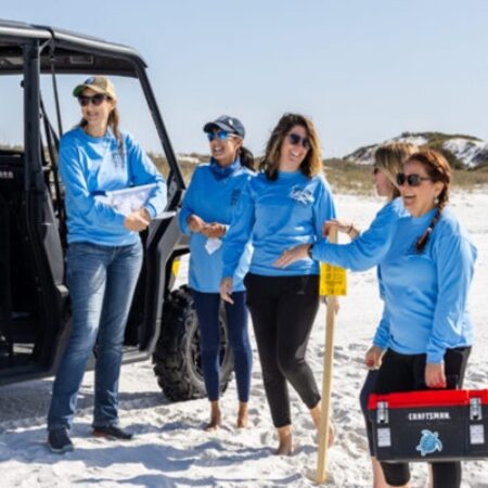 Five women in blue shirts stand on a sandy beach near a utility vehicle, holding tools and smiling, with dunes visible in the background.
