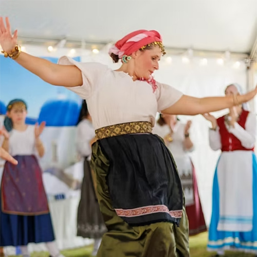 A woman in traditional clothing dances with arms outstretched in front of a group, also in folk attire, under a tent with string lights.