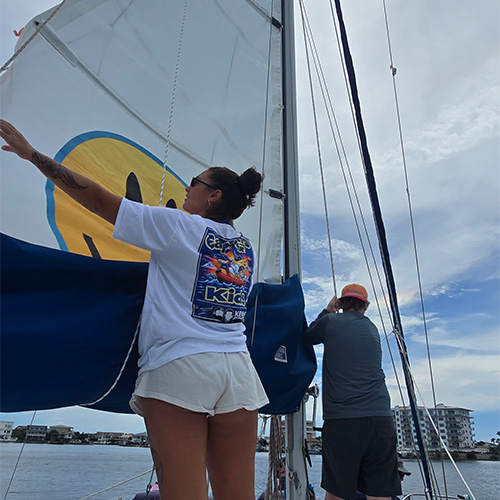 Two people adjust the sail on a boat; one faces the sail while the other looks ahead. Buildings and cloudy sky are visible in the background.